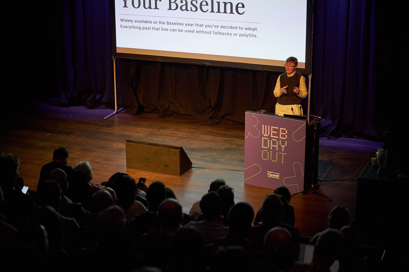 The stage at Web Day Out 2026, where Rachel Andrew is at the podium talking about Baseline and browser support. The podium is purple and features the text “Web Day Out”. Behind Rachel is a project showing her slides, and in front you can see the first few rows of the audience.