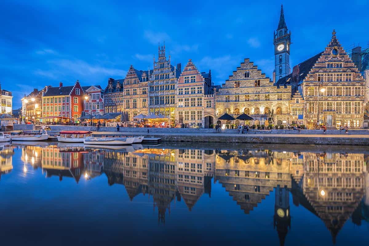 Night view of Ghent's waterfront, with illuminated historic architecture and a clock tower reflected in the canal